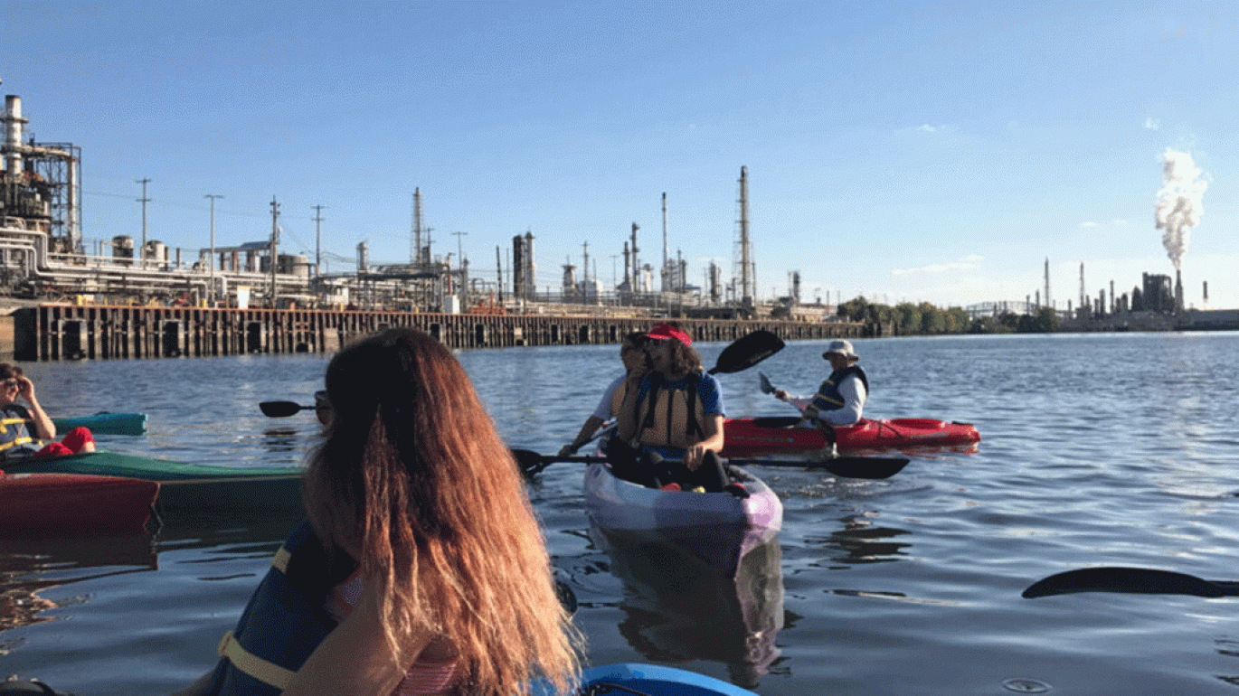 People kayaking on the water with an oil refinery visible in the background