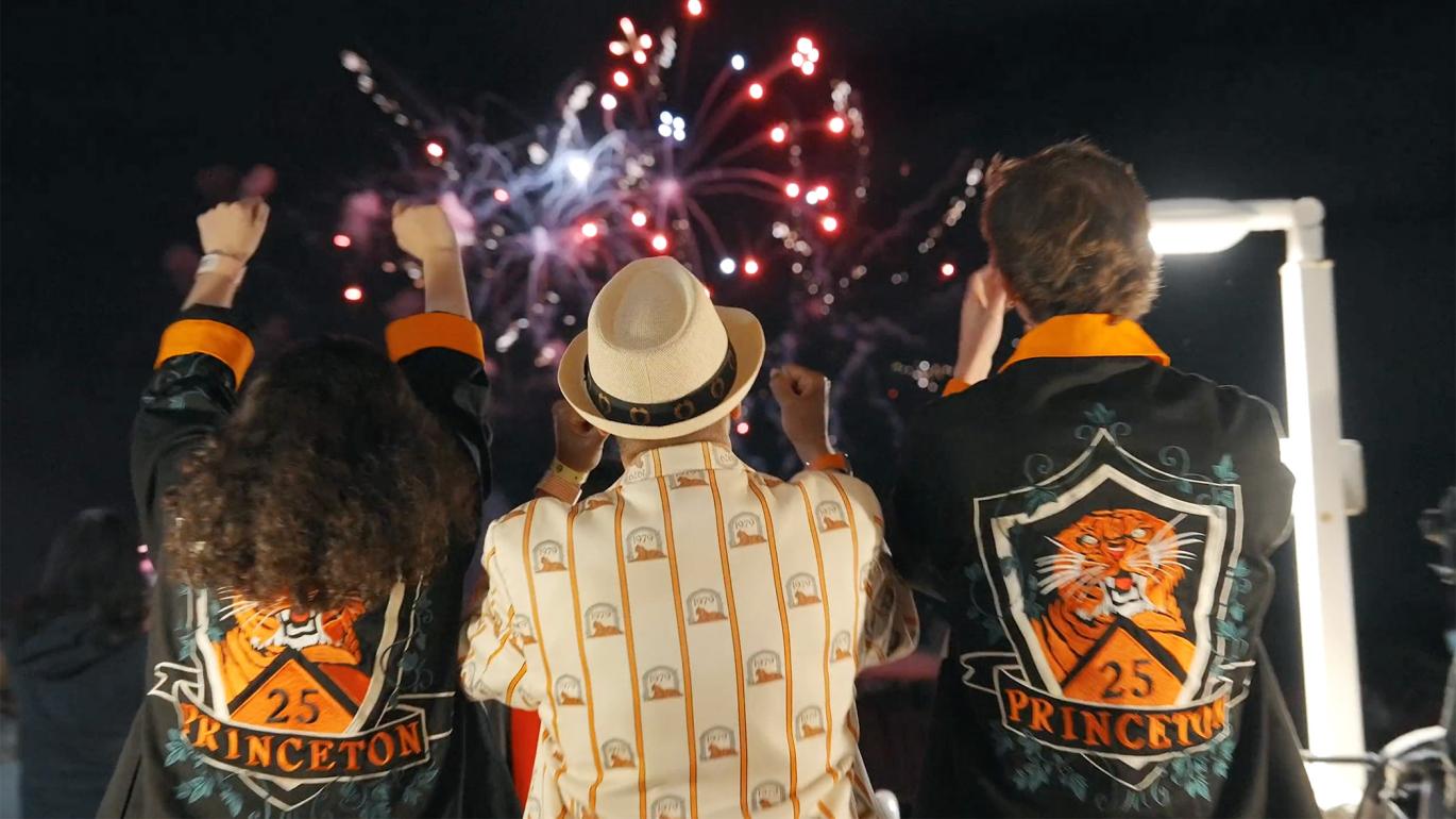 With fireworks in the background, three Princeton alumni, dressed in orange and black, cheer at Reunions. 