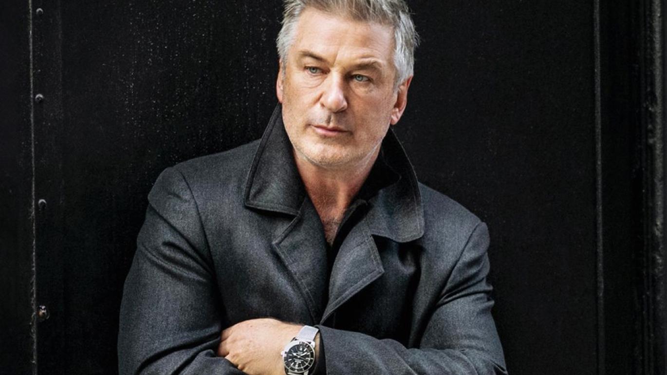 Alec Baldwin crosses his arms. He is wearing a black jacket.