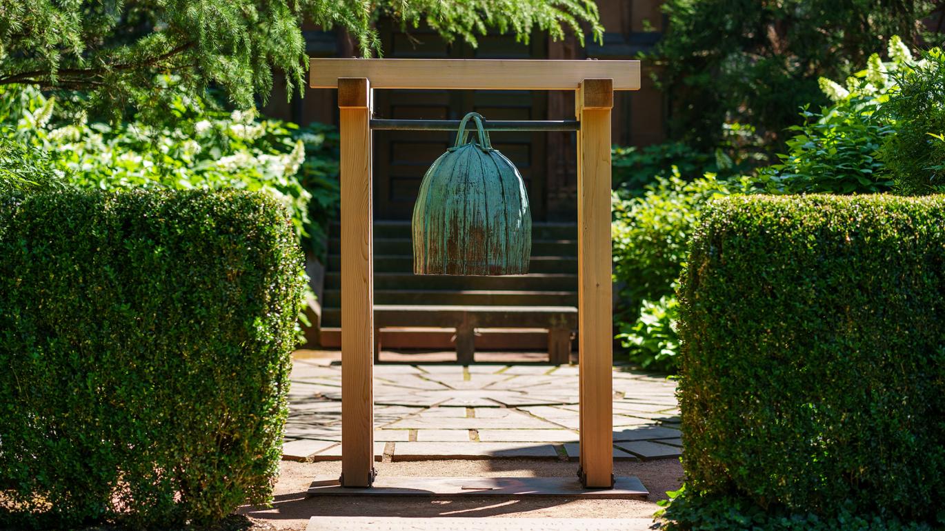 A greenish bell hanging fom a wooden post-frame in a garden