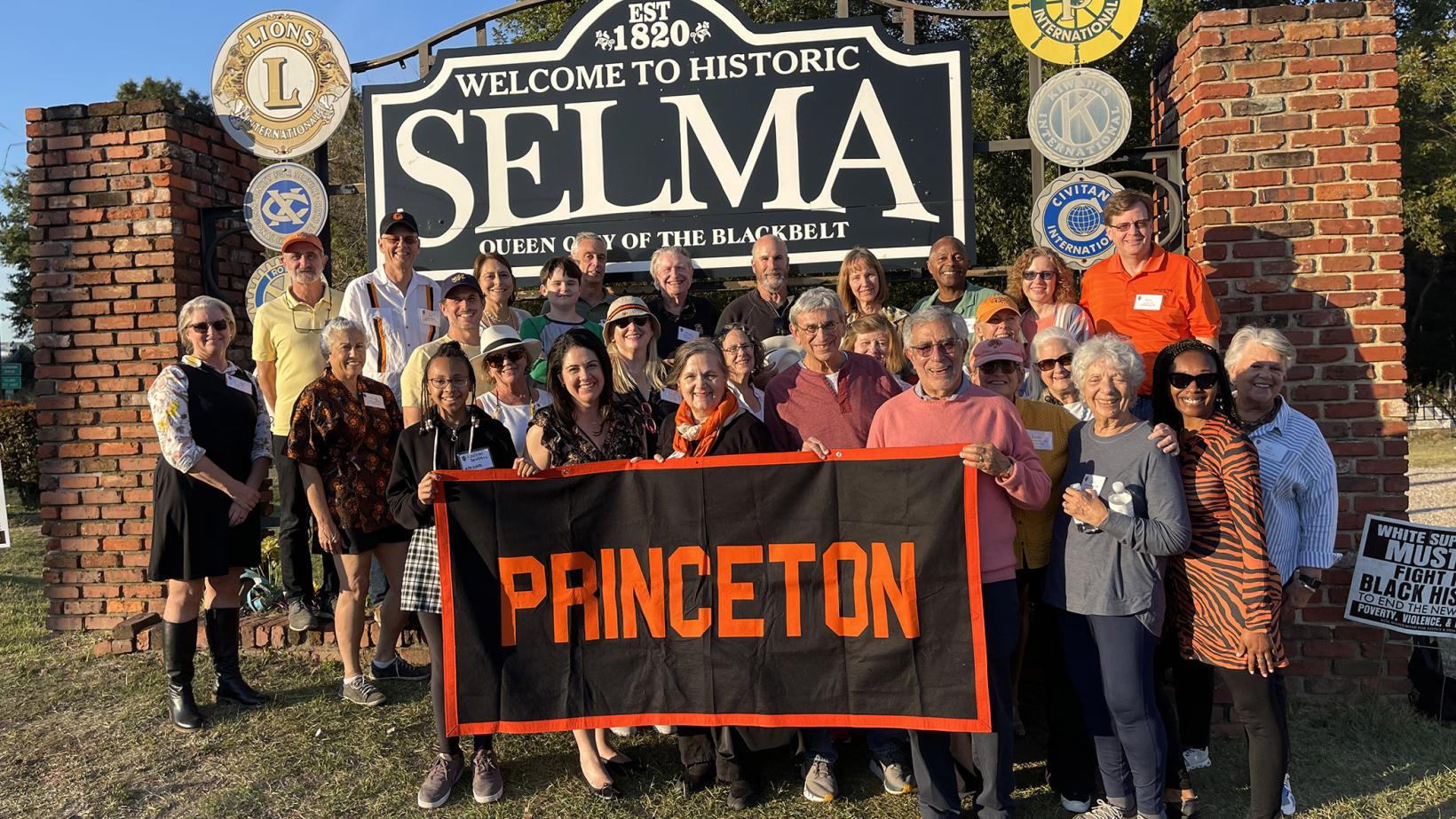 Princeton alumni and friends pose with a Princeton banner in front of a welcome sign to Selma, Alabama