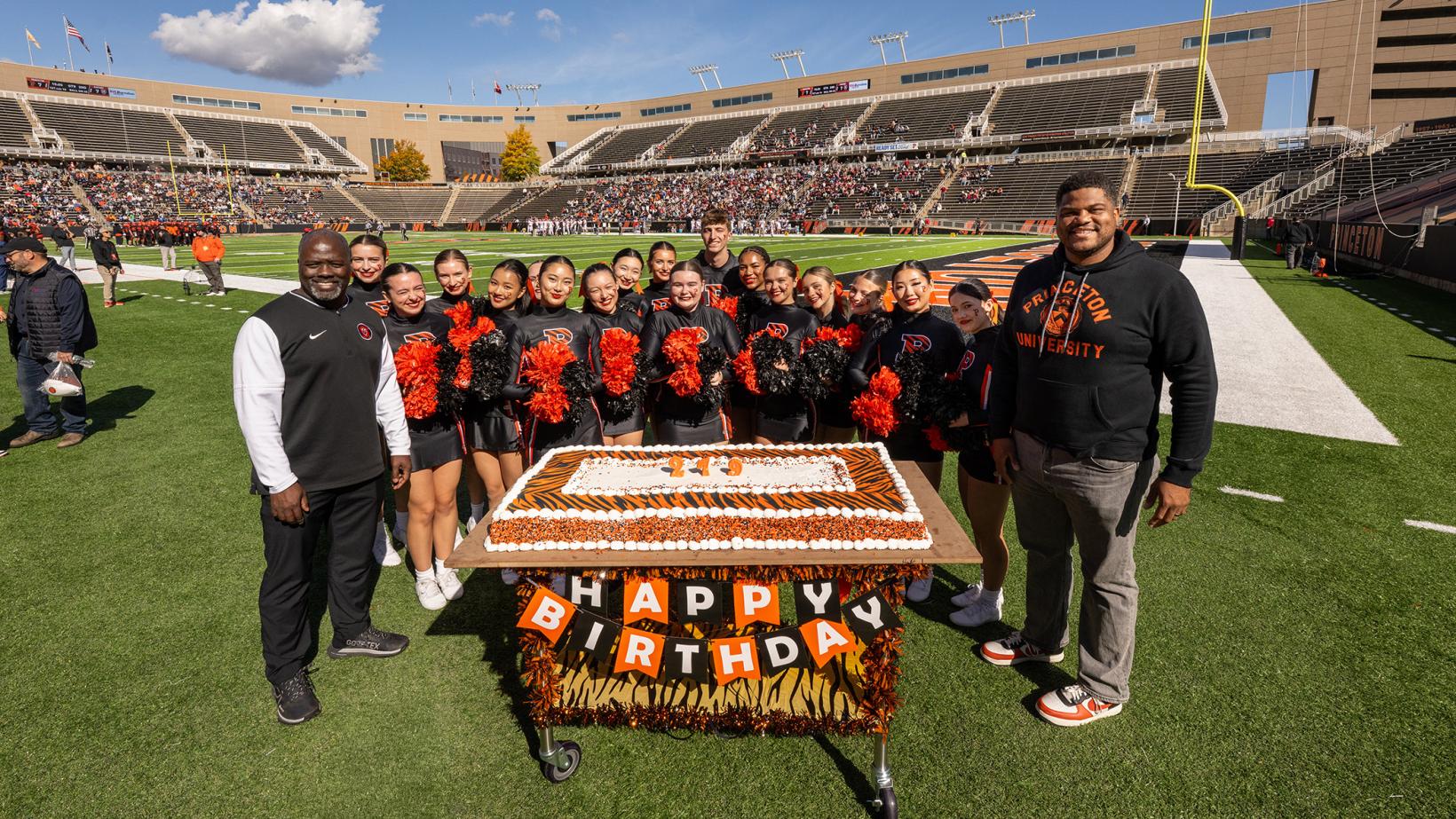 John Mack, Erik Plummer and the Princeton cheerleaders huddle around an orange birthday cake in Princeton Stadium
