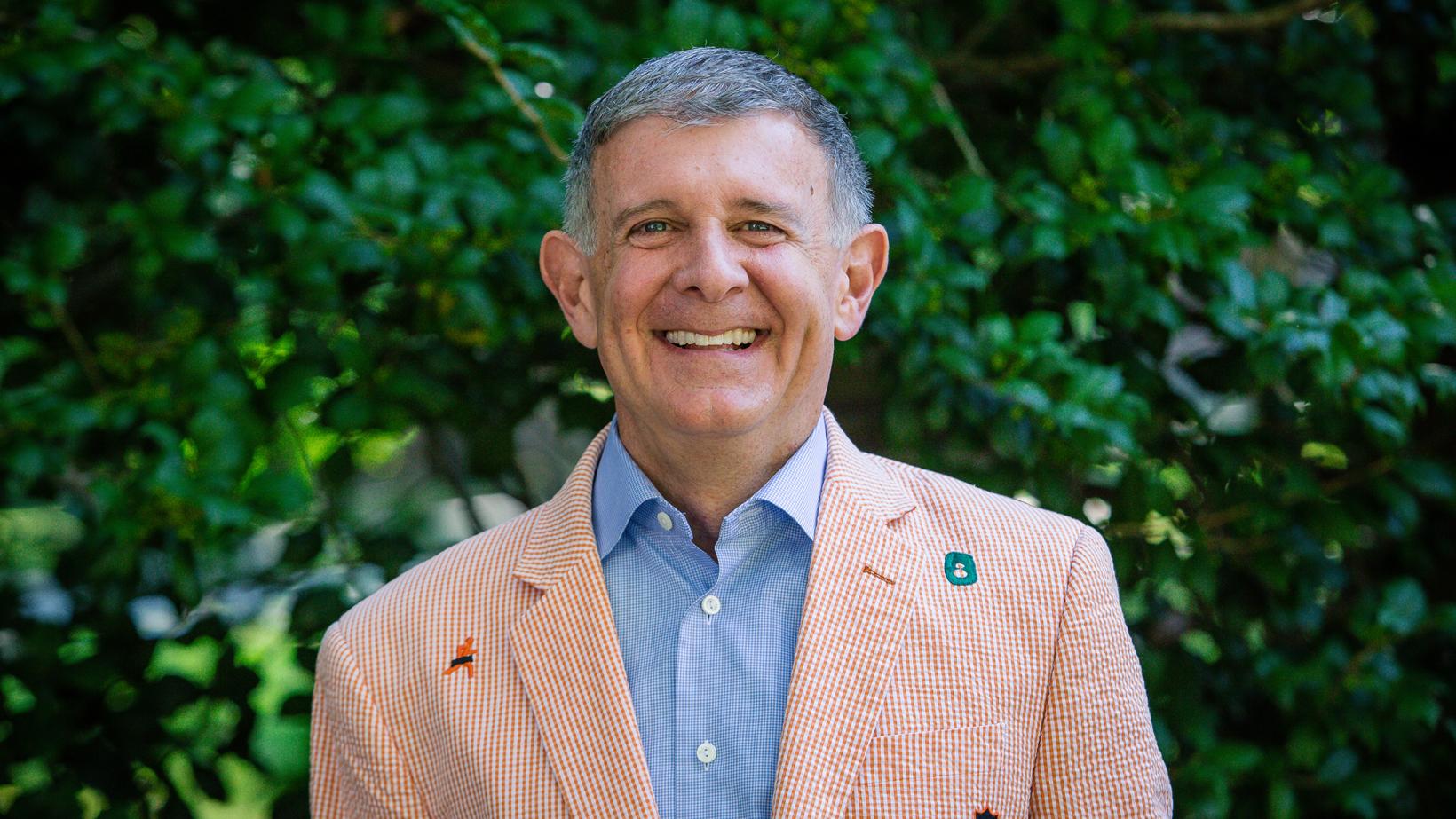 Man in a checkered blazer smiling in front of greenery.