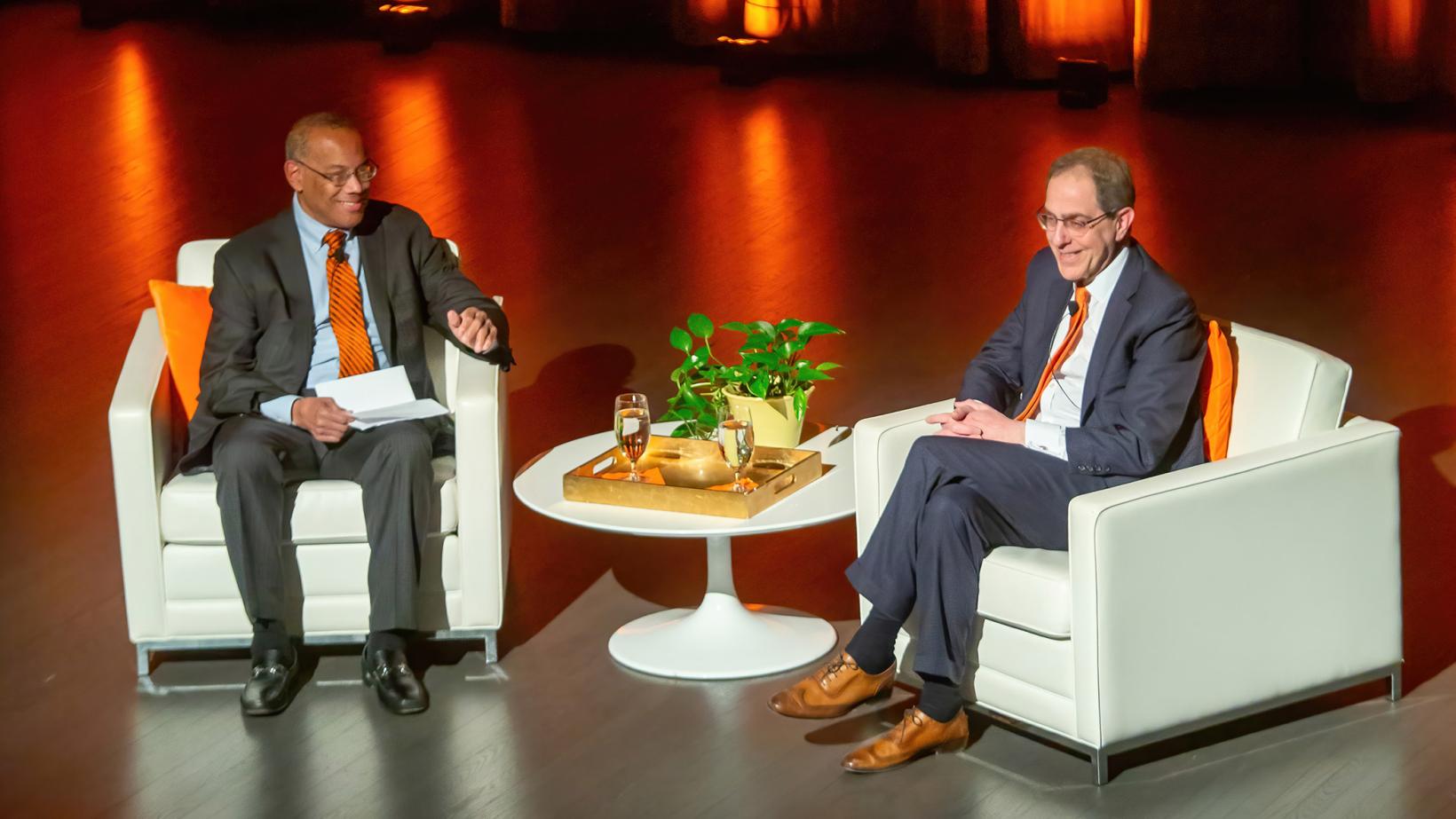 President Eisgruber and John Rogers sit on an orange-lit stage, smiling at an audience. 