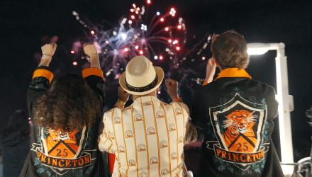 With fireworks in the background, three Princeton alumni, dressed in orange and black, cheer at Reunions. 