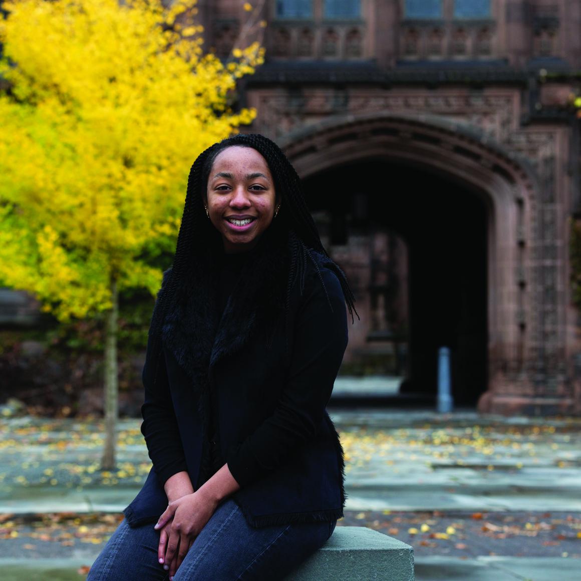Turquoise Brewington '22 seated on campus with stone archway and autumn tree in background