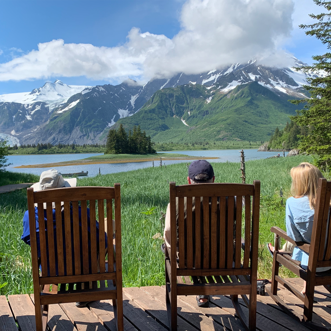 Back-view of travelers sitting in chairs enjoying view of Alaskan mountains.