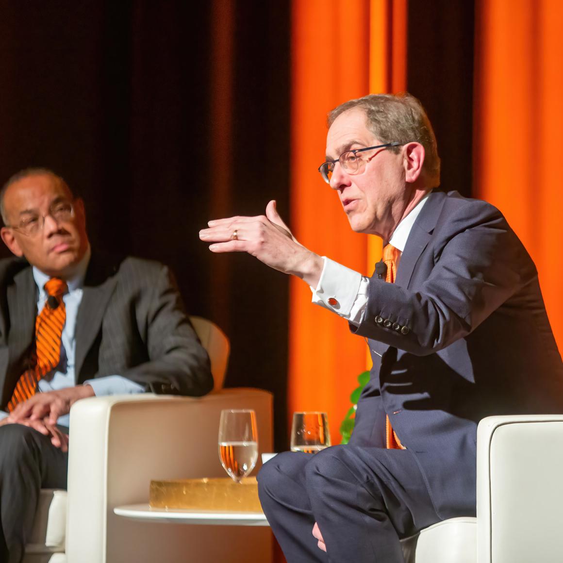 President Eisgruber motions with his left hand to make a point from the stage while John Rogers listens.
