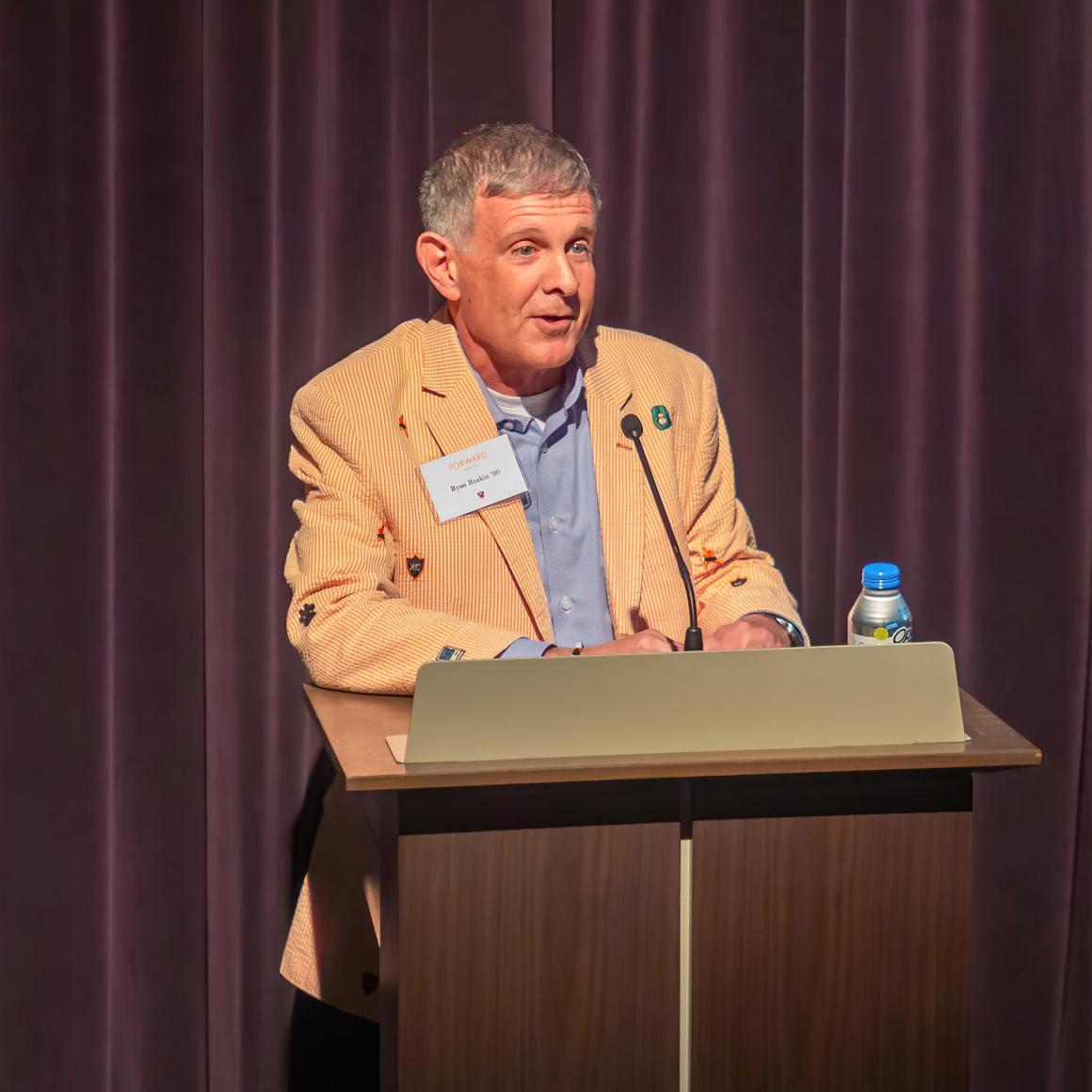 Ryan Ruskin, wearing his orange blzer, speaking to an audience from behind a lectern.