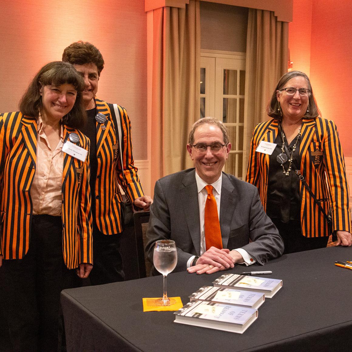 Christoper Eisgruber, seated a table with several of his books, surrounded by three alumna dressed in orange and black stripes