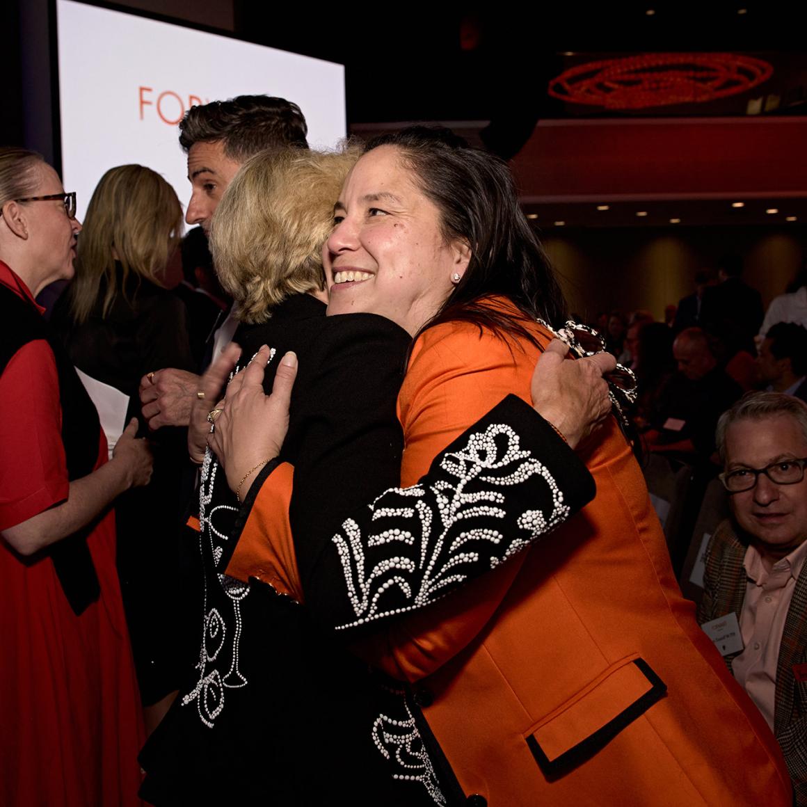 Two alumna wearing orange and black embracv warmly in a hall full of Princetonians.