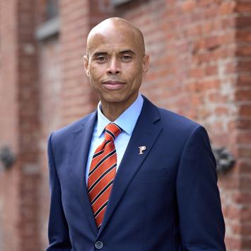 Laurence Latimer, wearing a blue suit and striped orange tie, standing near a brick building