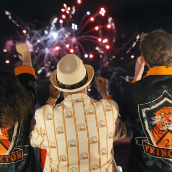 With fireworks in the background, three Princeton alumni, dressed in orange and black, cheer at Reunions. 