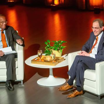 President Eisgruber and John Rogers sit on an orange-lit stage, smiling at an audience. 