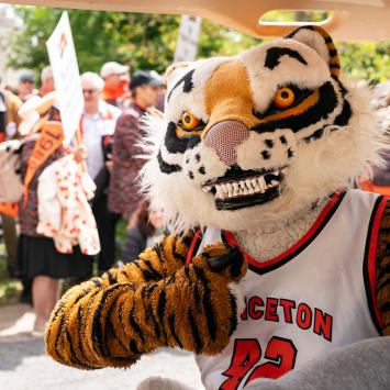 A person in a Tiger costume gives a thumbs up while riding in a golf cart; parade onlookers are in the background