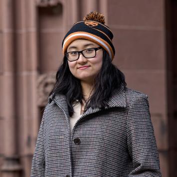 Virginia Jiang, wearing a black and organge knit cap, standing in front of a campus academic building.