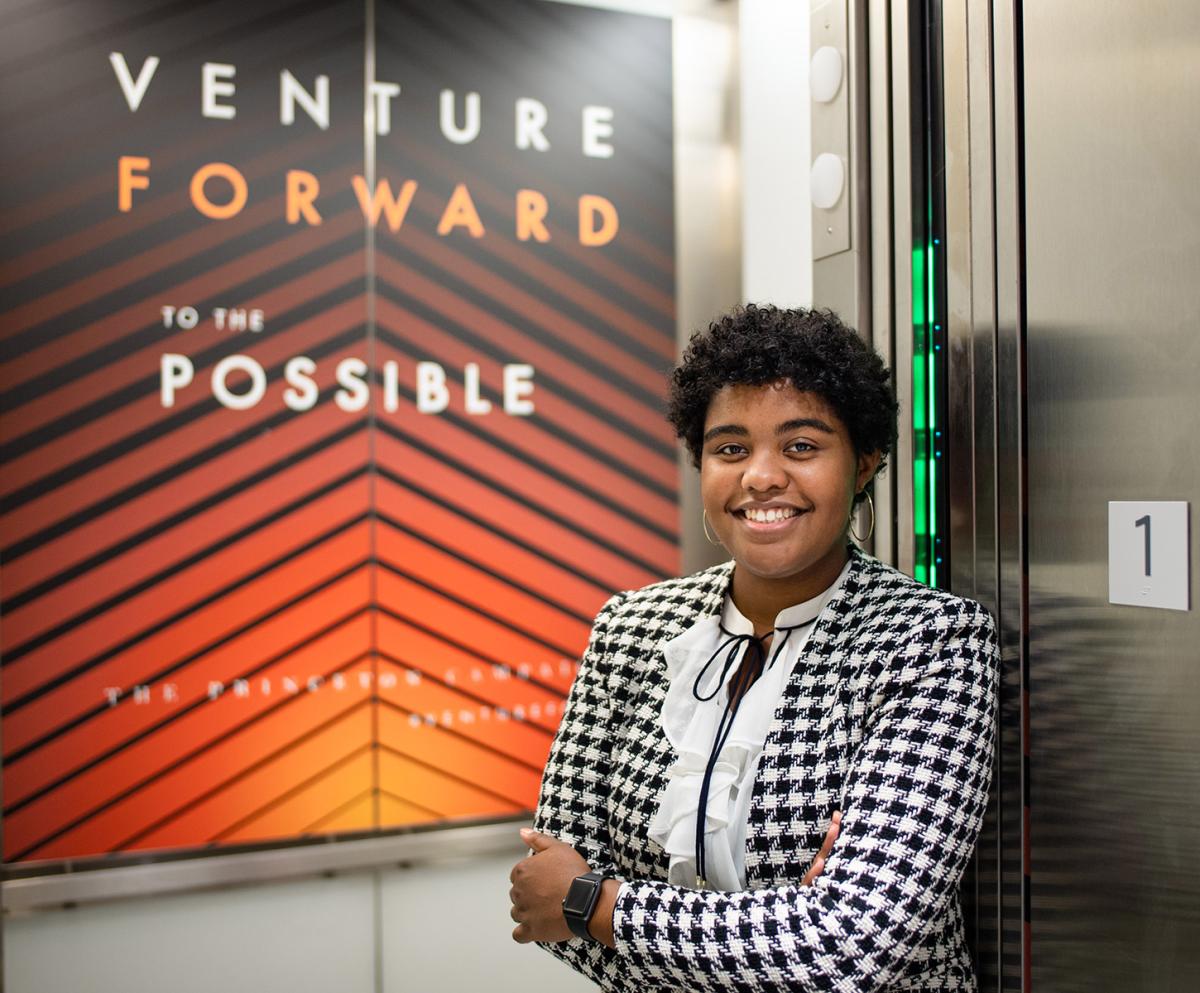 Morgan Smith, arms crossed and smiling, standing in front of an orange sign promoting the Venture Forward campaign