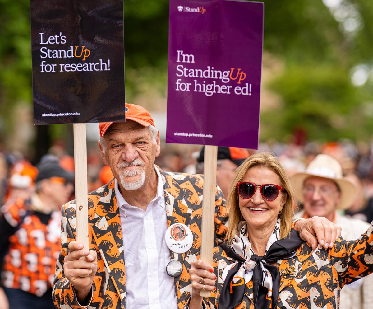 Two brightly-dressed alums in the P-rade hold signs supporting the Stand Up campaign for higher education.