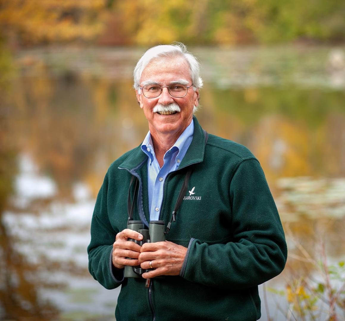 John Fitzpatrick stands in front of a body of water holding binoculars in his hands