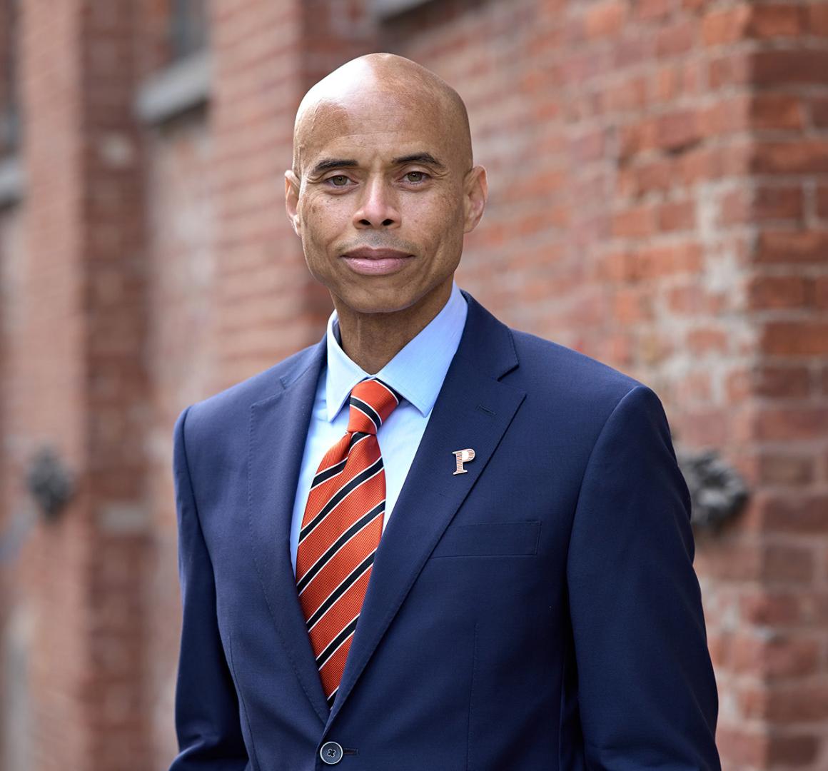 Laurence Latimer, wearing a blue suit and striped orange tie, standing near a brick building