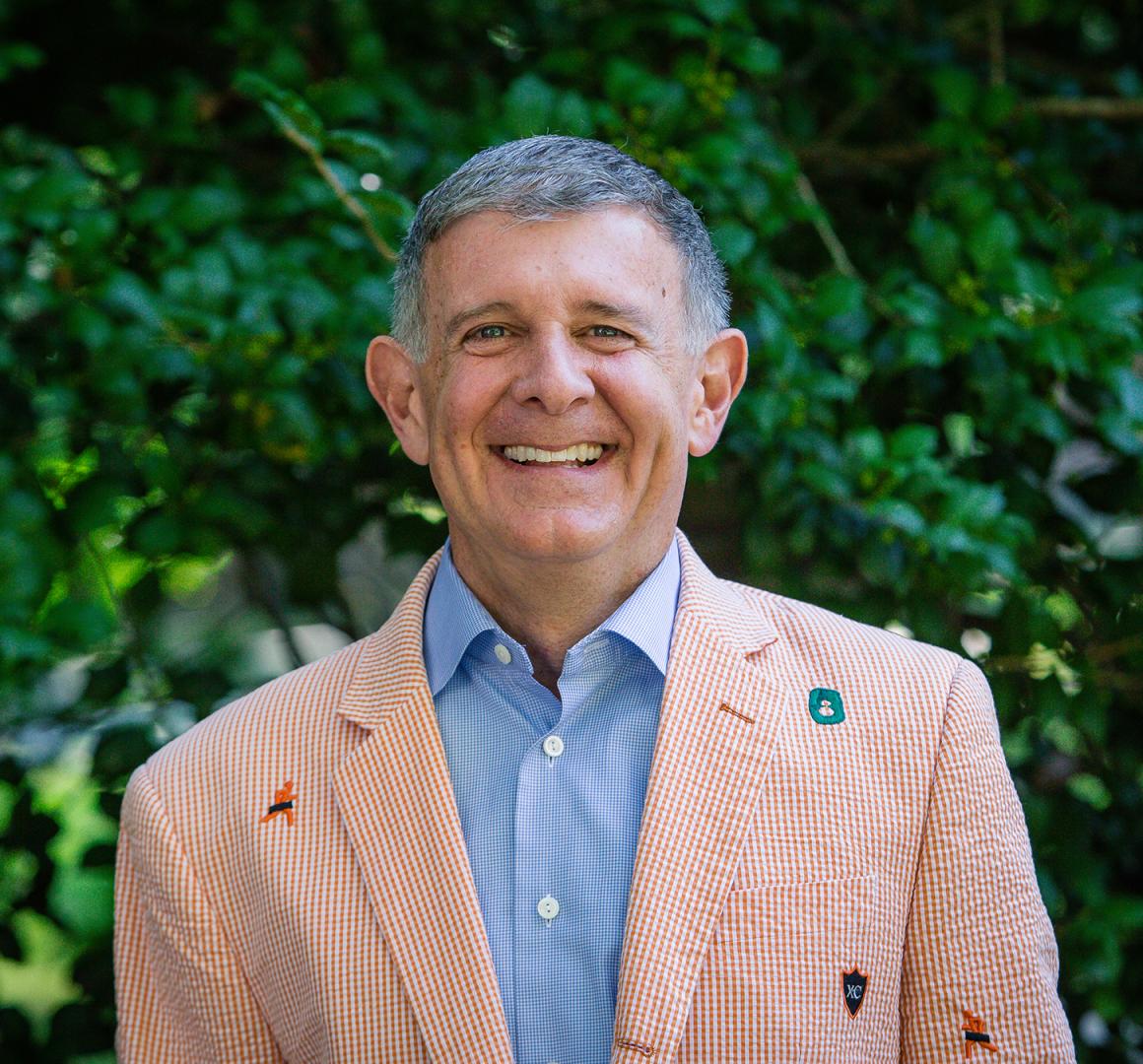 Man in a checkered blazer smiling in front of greenery.