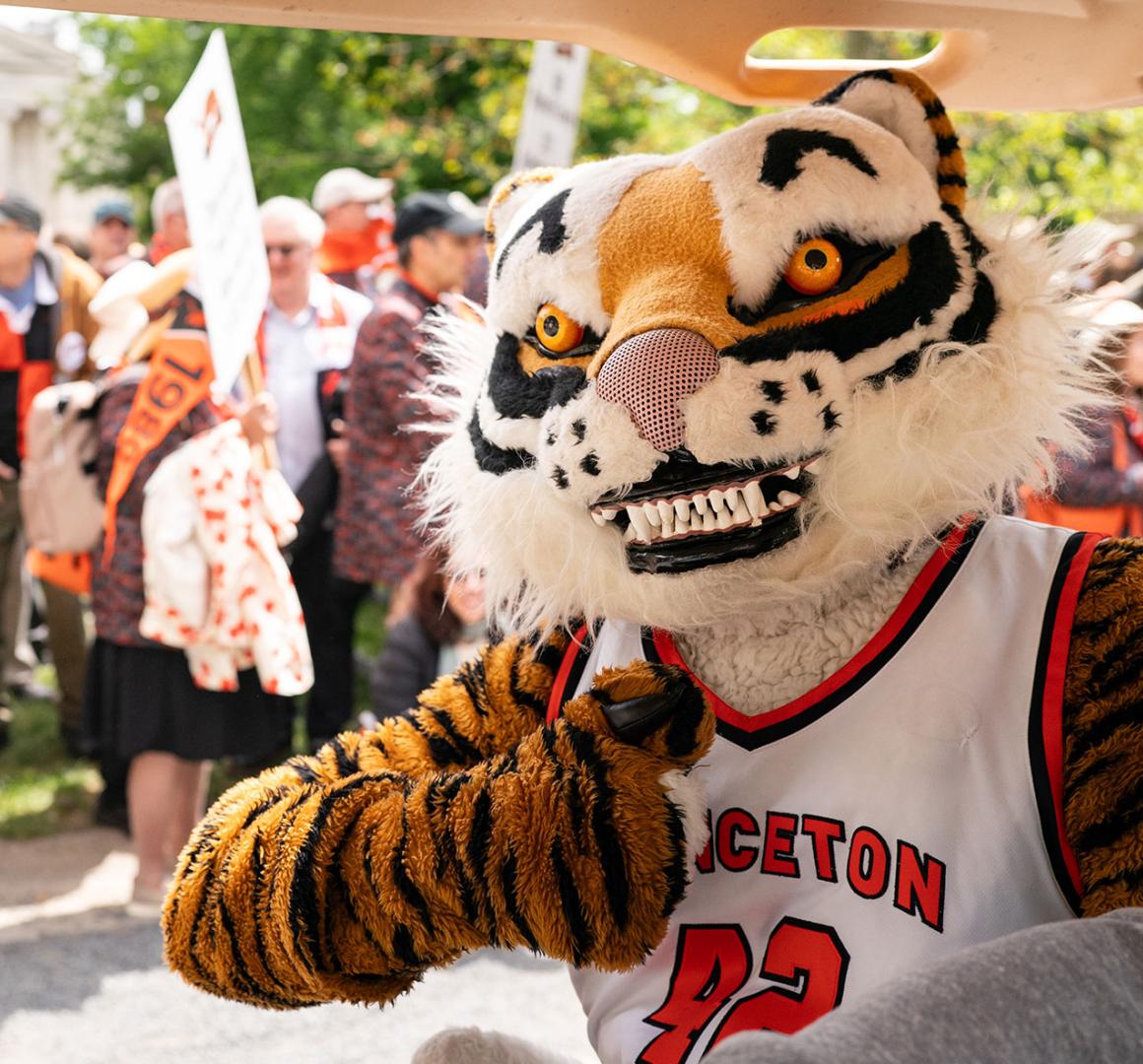 A person in a Tiger costume gives a thumbs up while riding in a golf cart; parade onlookers are in the background