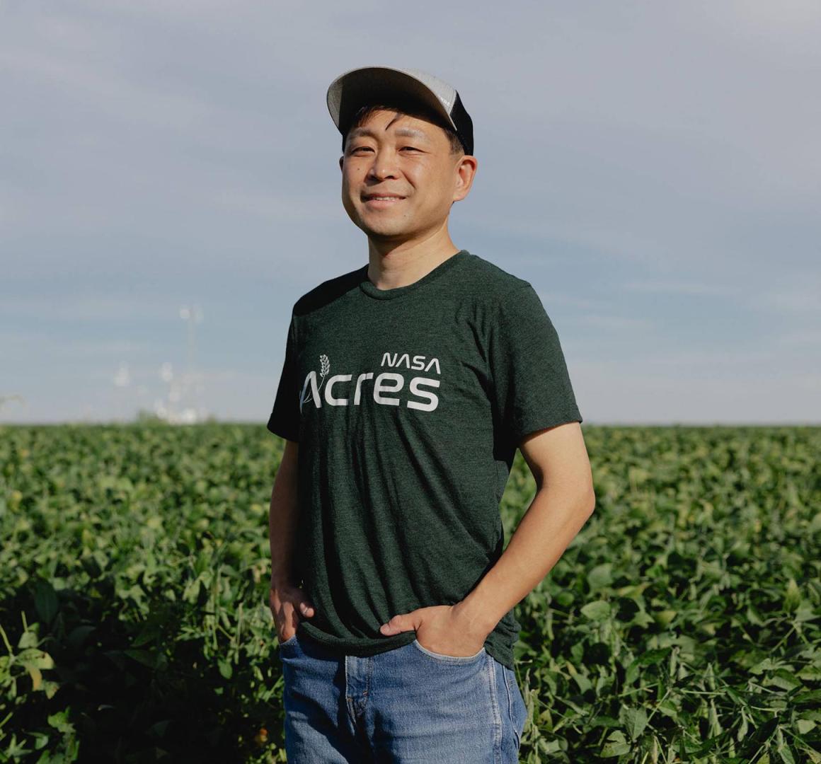 Asian male with baseball hat standing in a farm field with green crops behind him. 