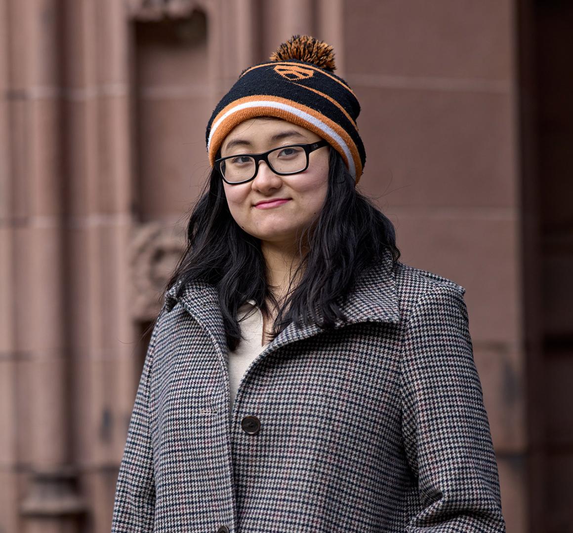 Virginia Jiang, wearing a black and organge knit cap, standing in front of a campus academic building.