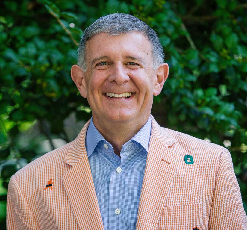 Man in a checkered blazer smiling in front of greenery.