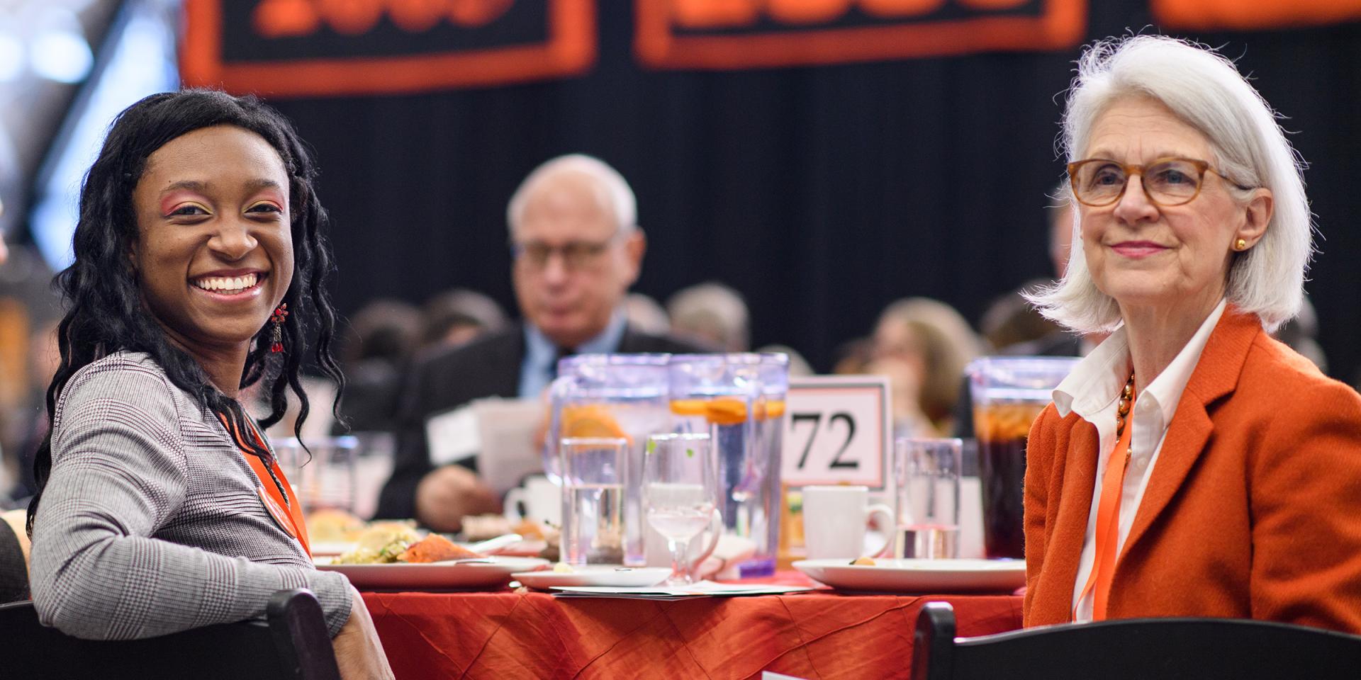 Two women sitting at table for Alumni Day with man in background.