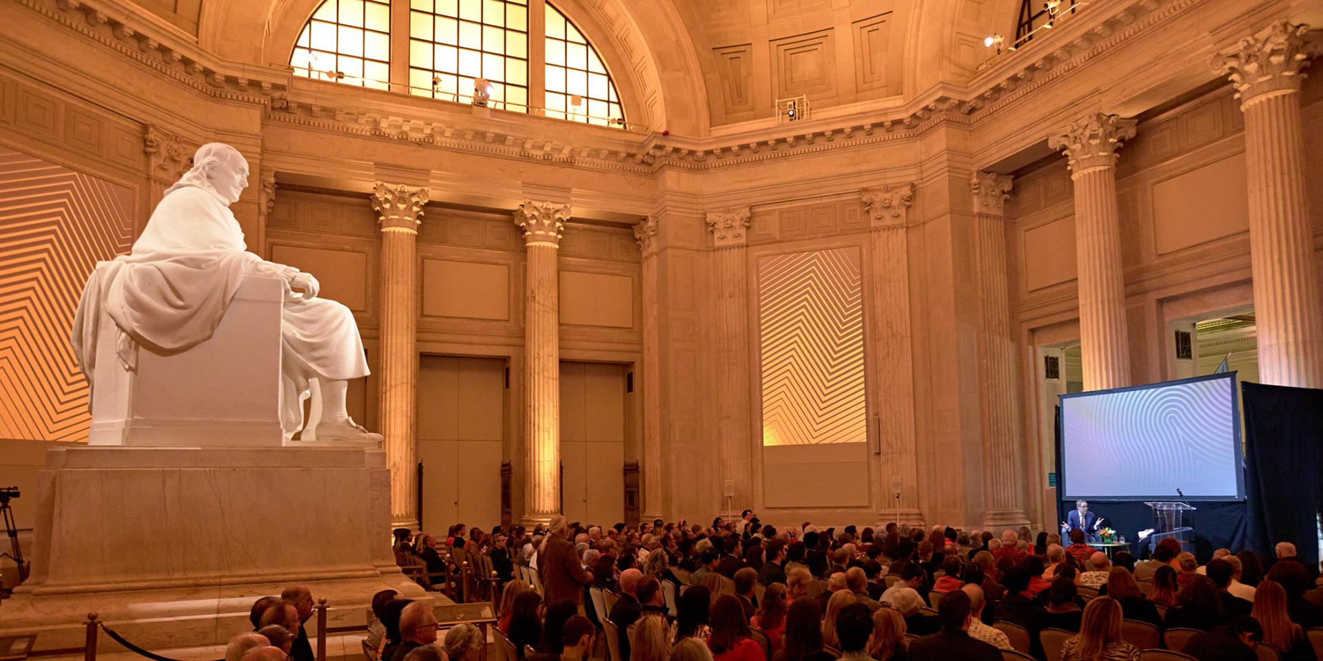 A statue of Benjamin Franklin looms over as crowd of Princeton alumni as they listen to President Eisgruber on stage at the Franklin Institute in Philadelphia. 