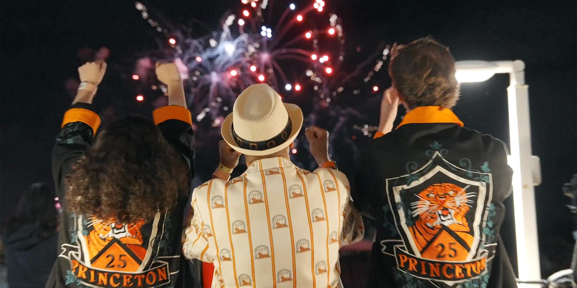 With fireworks in the background, three Princeton alumni, dressed in orange and black, cheer at Reunions. 