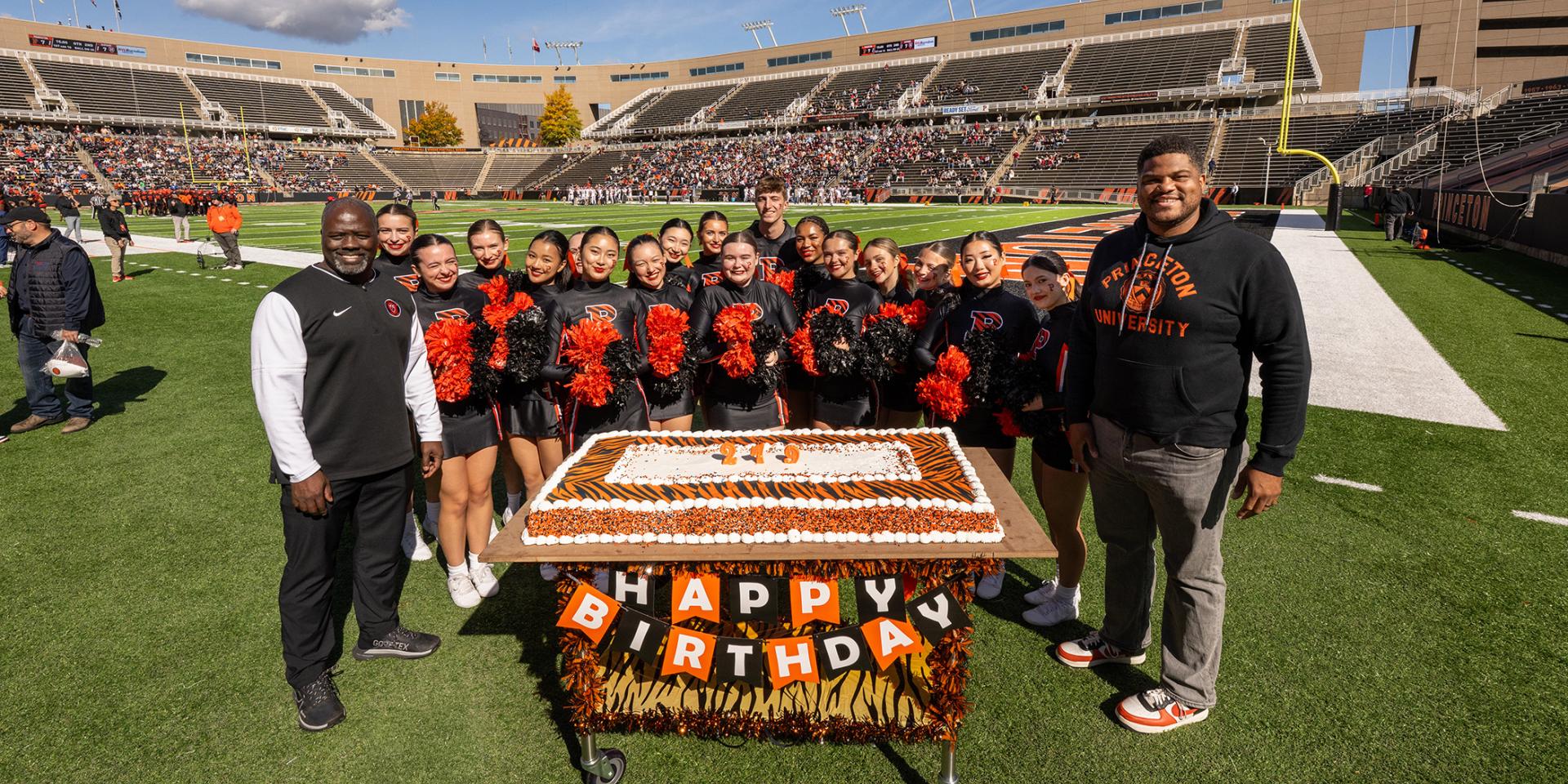 John Mack, Erik Plummer and the Princeton cheerleaders huddle around an orange birthday cake in Princeton Stadium