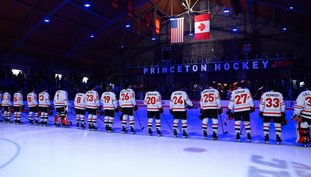 The Princeton men's ice hockey team, wearing white jerseys, stands shoulder to shoulder during the national anthem. 