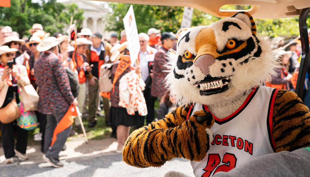 A person in a Tiger costume gives a thumbs up while riding in a golf cart; parade onlookers are in the background