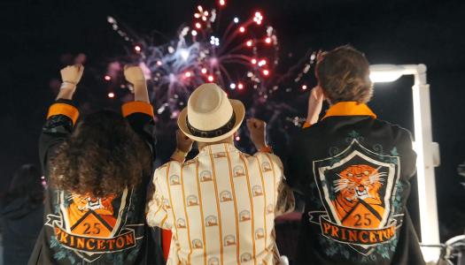 With fireworks in the background, three Princeton alumni, dressed in orange and black, cheer at Reunions.