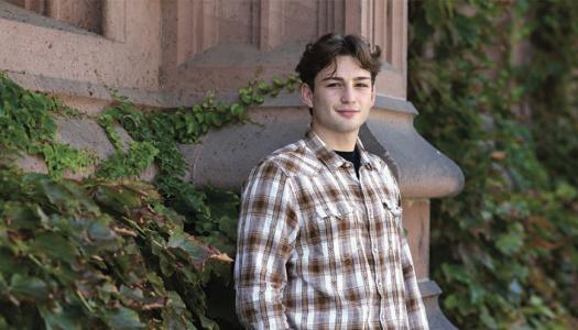 Male Princeton stone leans against the ivy-covered side of a campus building. 