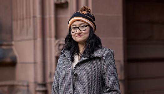 Virginia Jiang, wearing a black and organge knit cap, standing in front of a campus academic building.