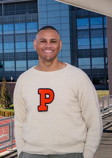 A man wearing a white sweater with an orange letter P on it stands in front of an outdoor track