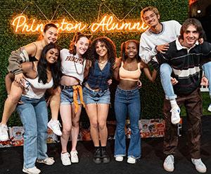 Seven young alumni pose in front of a neon Princeton Alumni sign