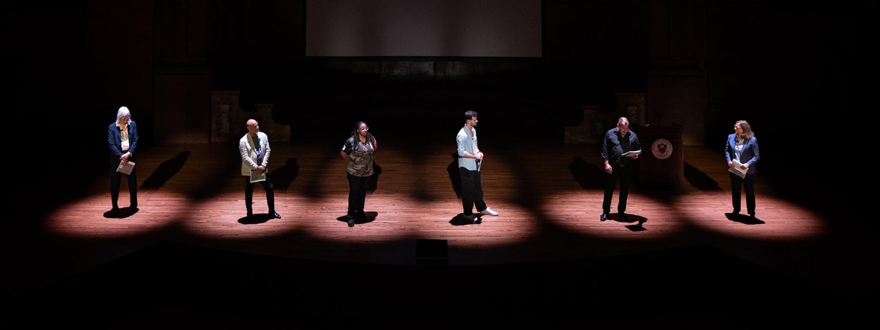 Six alumni stand in six spotlights on the Richardson Auditorium stage