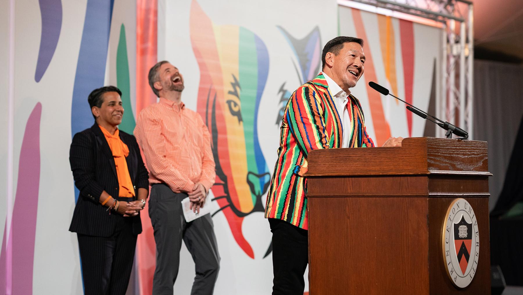 Timothy Wu speaks at the podium while Marisa Demeo and Brian Johnson look on and laugh