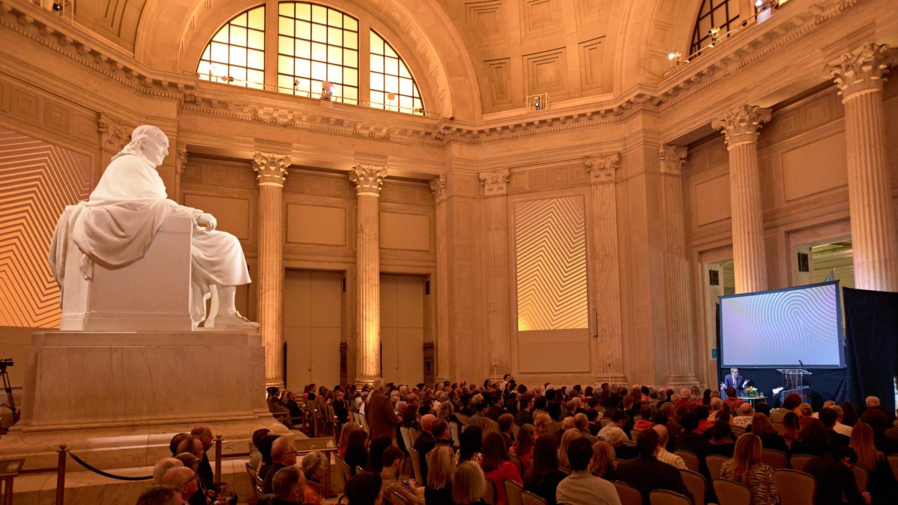 A statue of Benjamin Franklin looms over as crowd of Princeton alumni as they listen to President Eisgruber on stage at the Franklin Institute in Philadelphia.