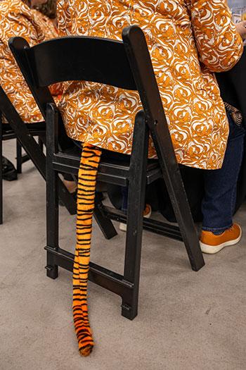 Back of a person seated in a chair with a tiger tail extending from the bottom of his jacket