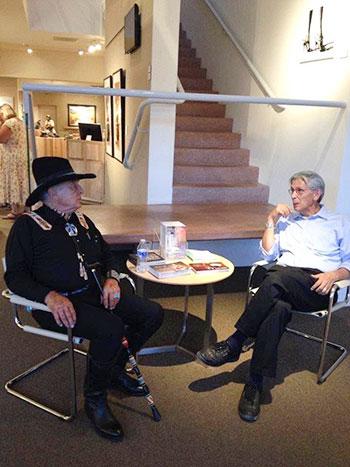 Two men sit at a table, looking at each other. The one at left, Ben Campbell, wears a hat and is holding a cane.