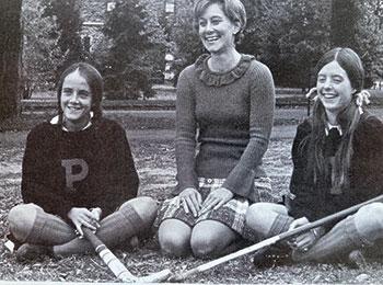 Coach Merrily Baker sits on the ground with two students on either side of her, both wearing Varsity sweaters and holding field hockey sticks; Bonnie Hewson is at right